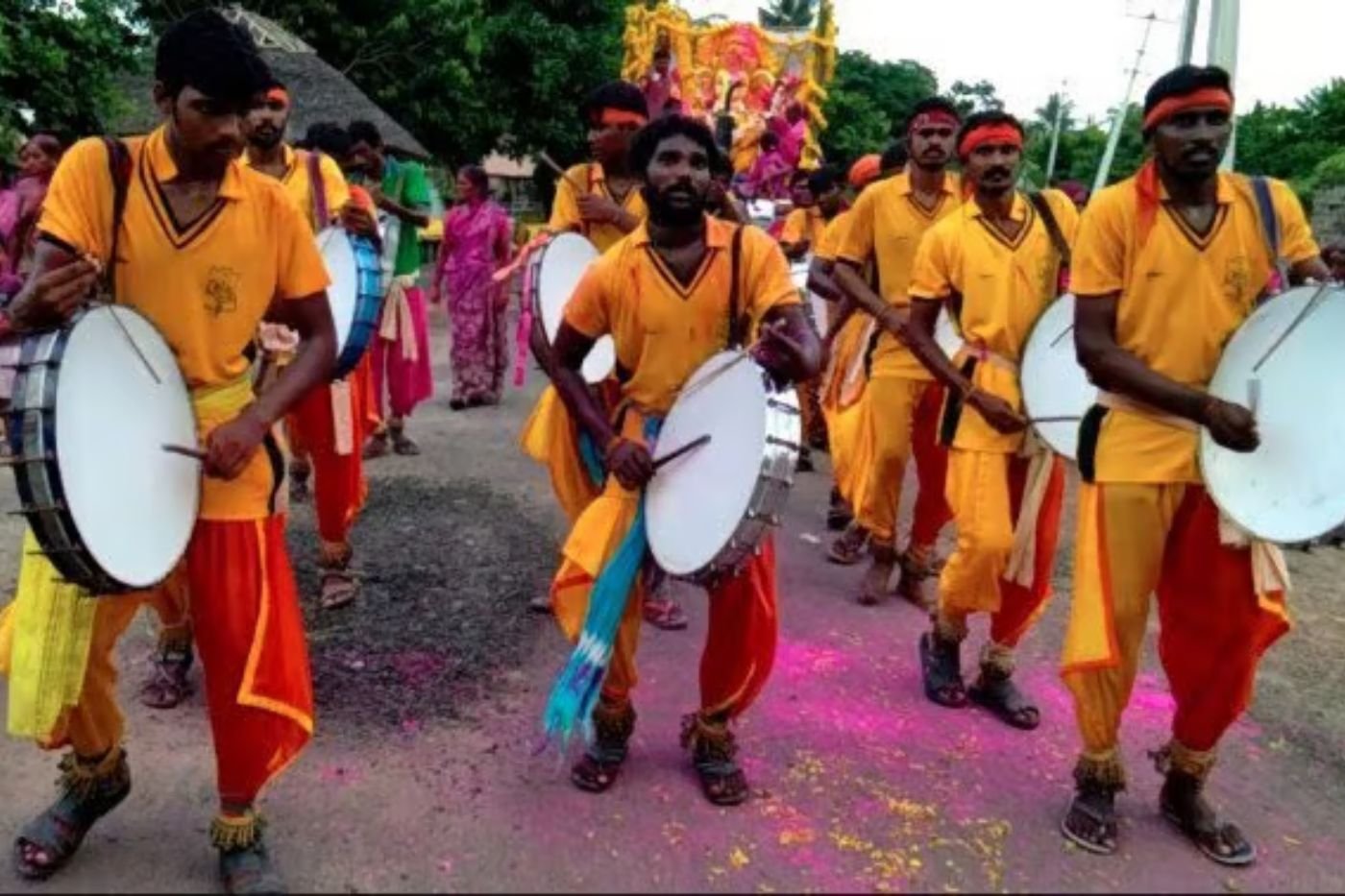 Dappu performers in yellow and orange costumes and headbands walking in a village procession, carrying round dappu drums and playing them with sticks