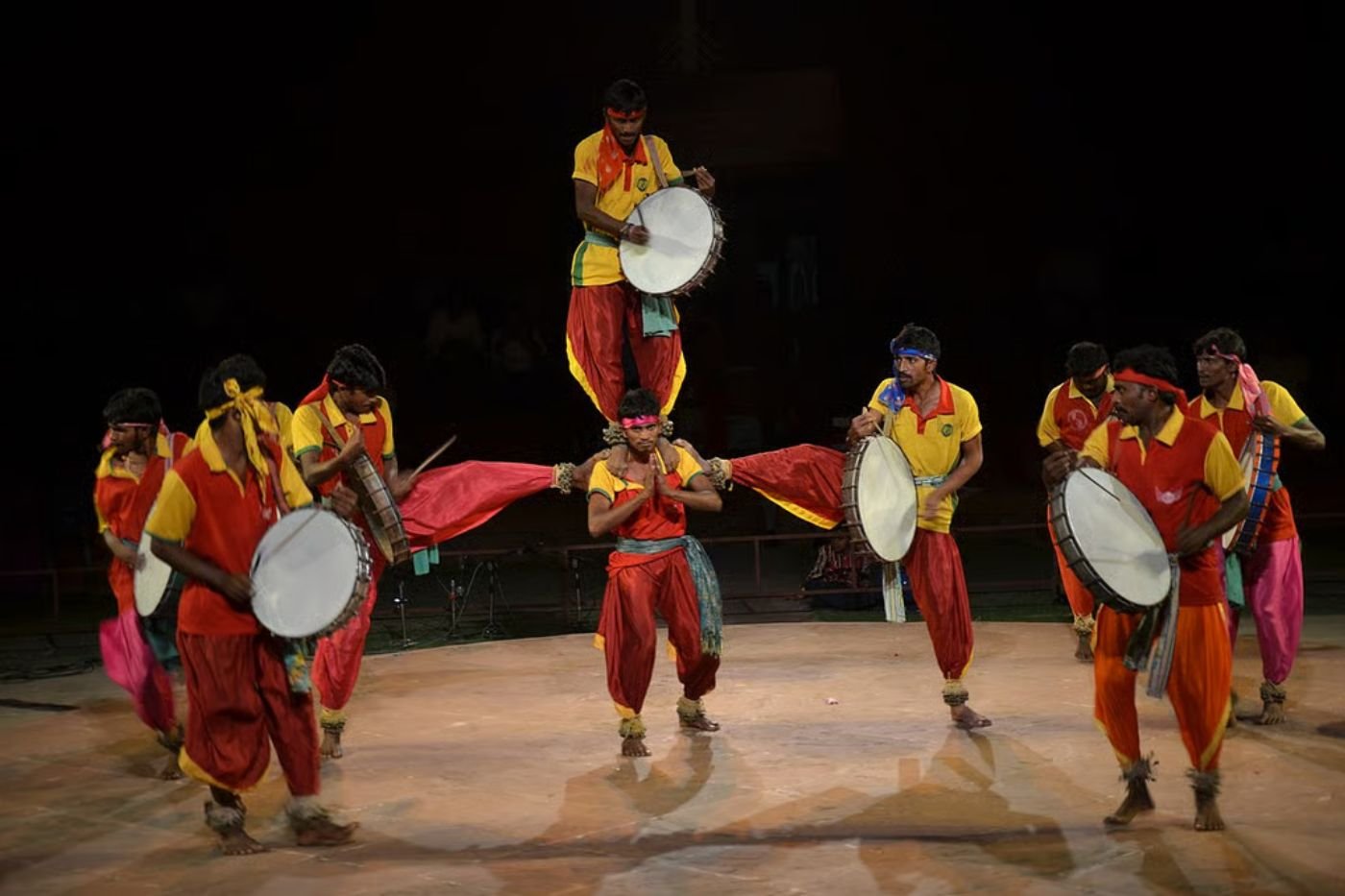 Dappu dancers on a floodlit night stage in yellow and red shirts and ankle bells, playing frame drums while two performers form a human pyramid