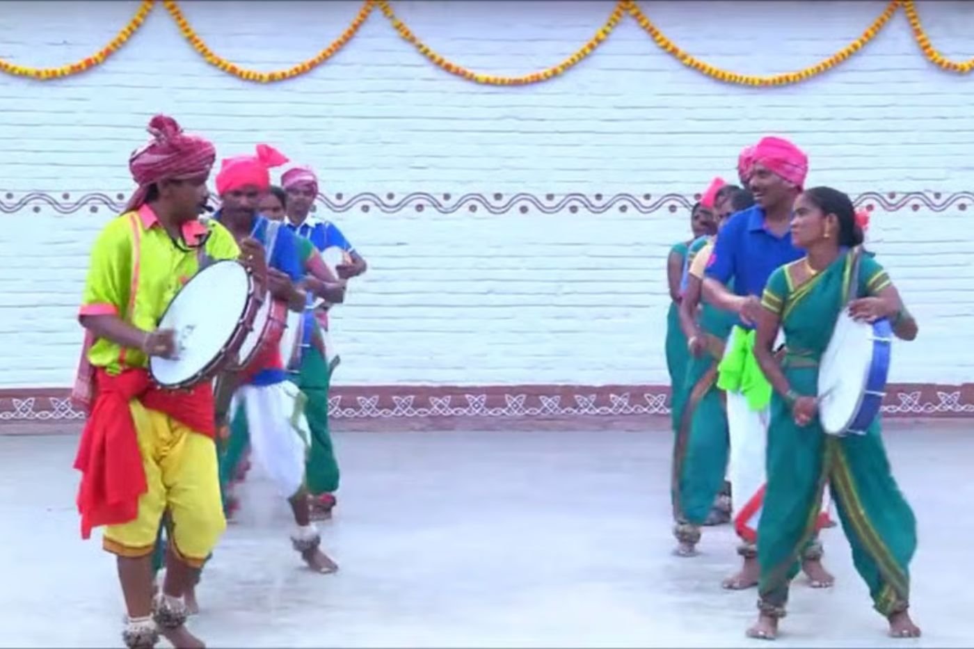 Dappu group in colourful turbans, dhotis and saris holding round frame drums during an outdoor performance in front of a decorated courtyard wall
