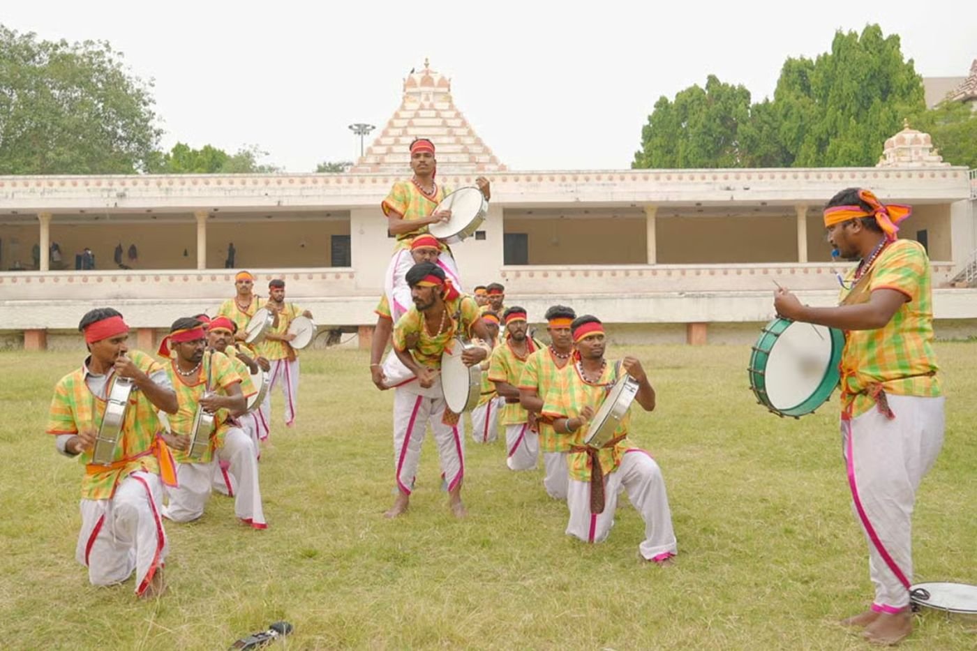 Dappu dancers in checked shirts, white dhotis and headbands playing large round frame drums with sticks, one performer lifted onto a teammate's shoulders outside a temple-style building